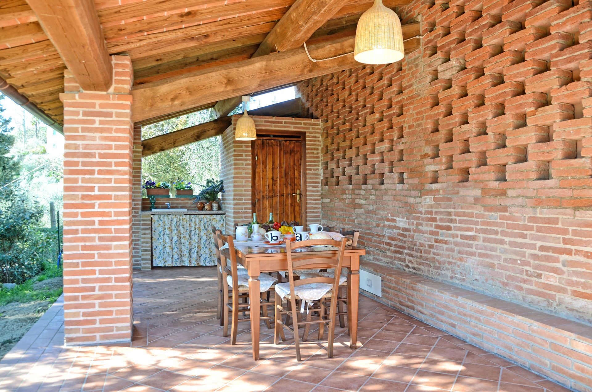 Une terrasse couverte avec un mur en briques, des poutres en bois au plafond et une table dressée pour le repas.