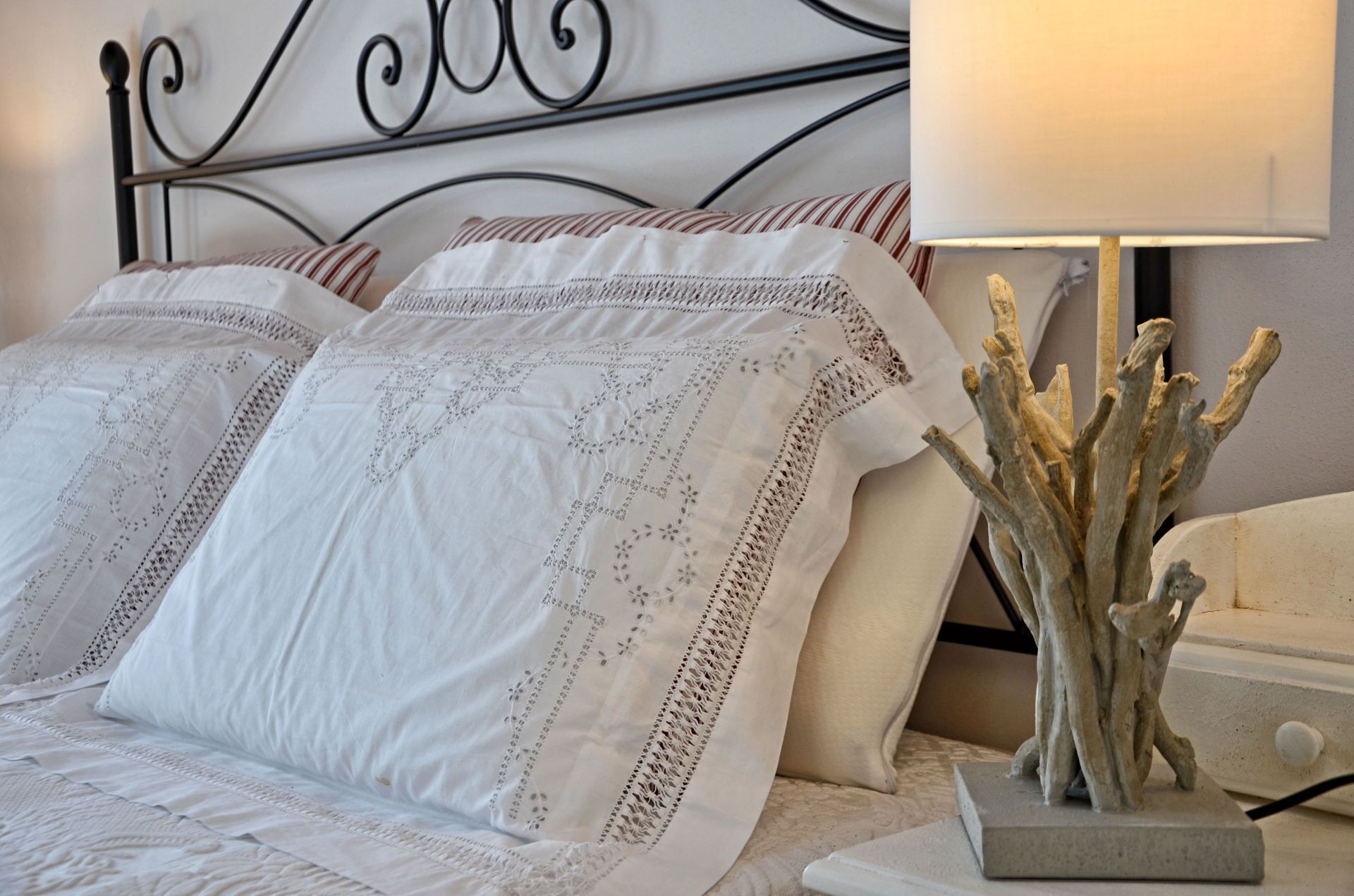 Ein ordentlich bezogenes Bett mit weißen Leinen und dekorativen Kissen, ein Metall-Kopfteil sowie ein Nachttisch mit Lampe und Zimmerpflanze.