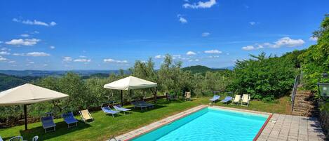 A pool area with lounge chairs and umbrellas, surrounded by greenery and a view of distant hills.