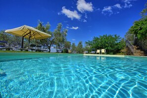 Une piscine aux eaux cristallines, bordée de chaises longues et d’ombrelles, entourée d’arbres et d’un escalier en pierre.