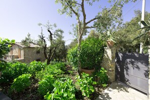 A stone house with a tiled roof, surrounded by lush greenery and a metal gate.