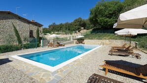 A rectangular swimming pool with a stone border, surrounded by lounge chairs and umbrellas, set against a backdrop of stone buildings and greenery.