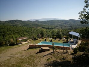 Un espace piscine doté de chaises longues et d’une structure à toit de chaume, entouré d’une végétation luxuriante et avec des collines en arrière-plan.