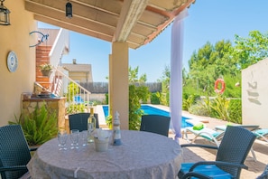 A covered outdoor dining area with a table set for four, surrounded by chairs, overlooking a swimming pool and greenery.