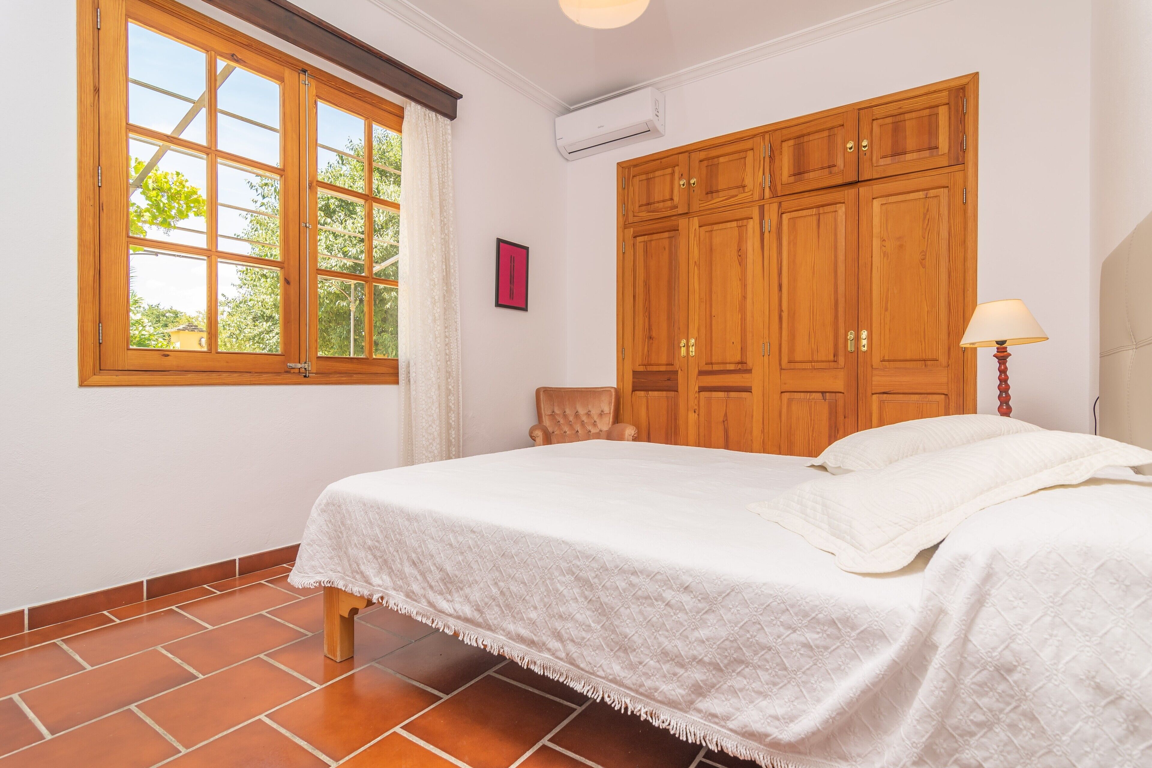 Une chambre avec une grande armoire en bois, un lit avec une literie blanche, une fenêtre avec des rideaux et une petite table avec une chaise.