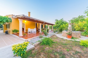 A yellow villa with a tiled patio, outdoor seating, and a garden with potted plants and flowers.