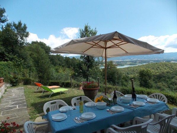 An outdoor dining area with a table set for six, surrounded by white chairs, under a large umbrella.