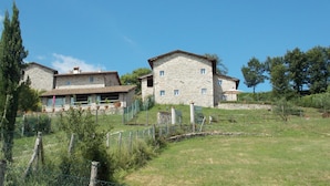 A stone building with multiple windows and a flat roof, surrounded by a fenced garden and trees.