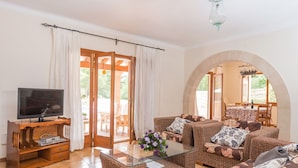 A living room with wicker furniture, a glass coffee table, a flat-screen TV, and a view through an arched doorway to an outdoor patio. - ES TURO DE FRANÇA - Villa with private pool in S'Horta. Free WiFi (Felanitx)