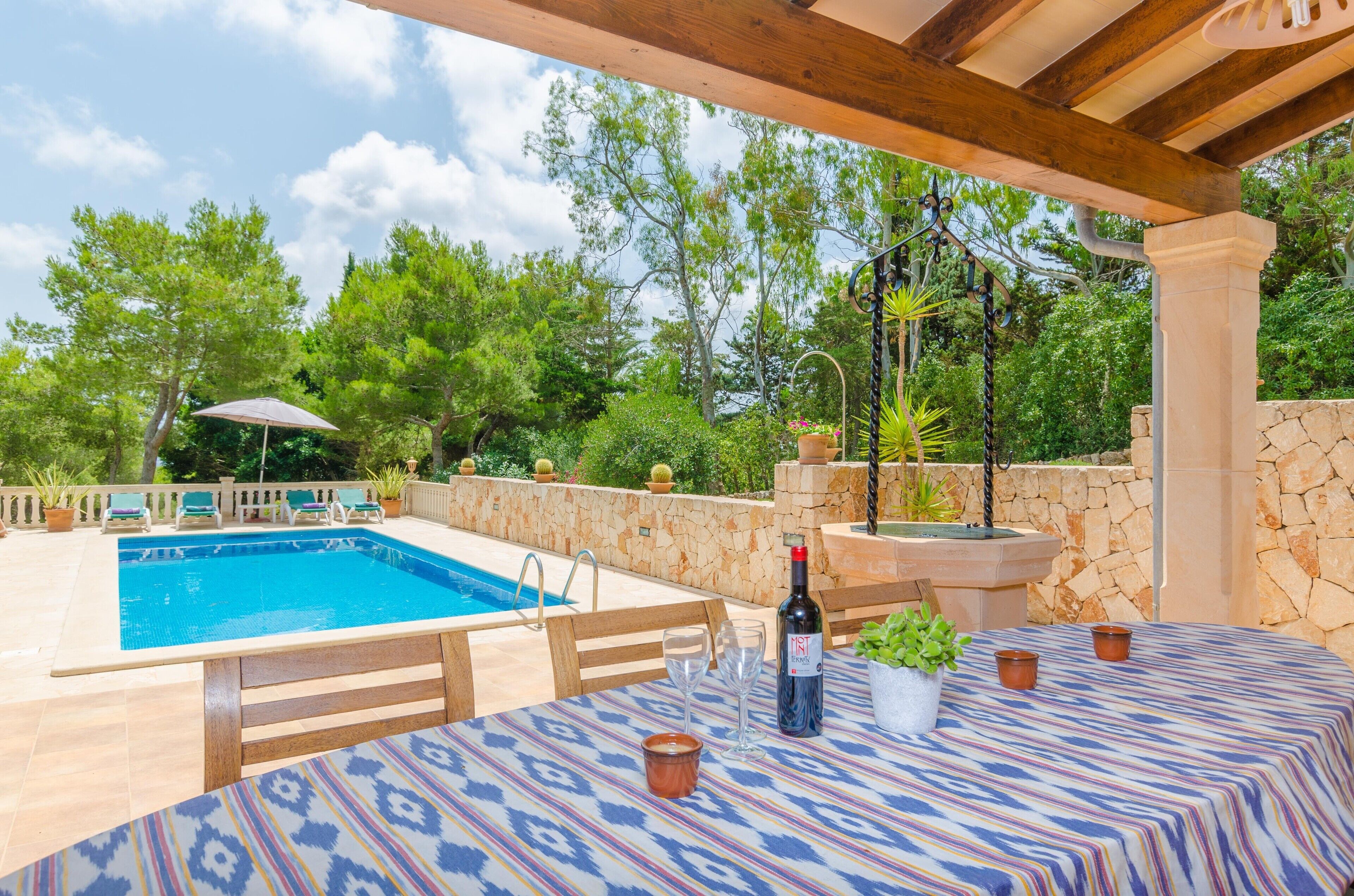 Area piscina esterna con un pergolato in legno, un tavolo con tovaglia fantasia, una bottiglia di vino e piante in vaso.