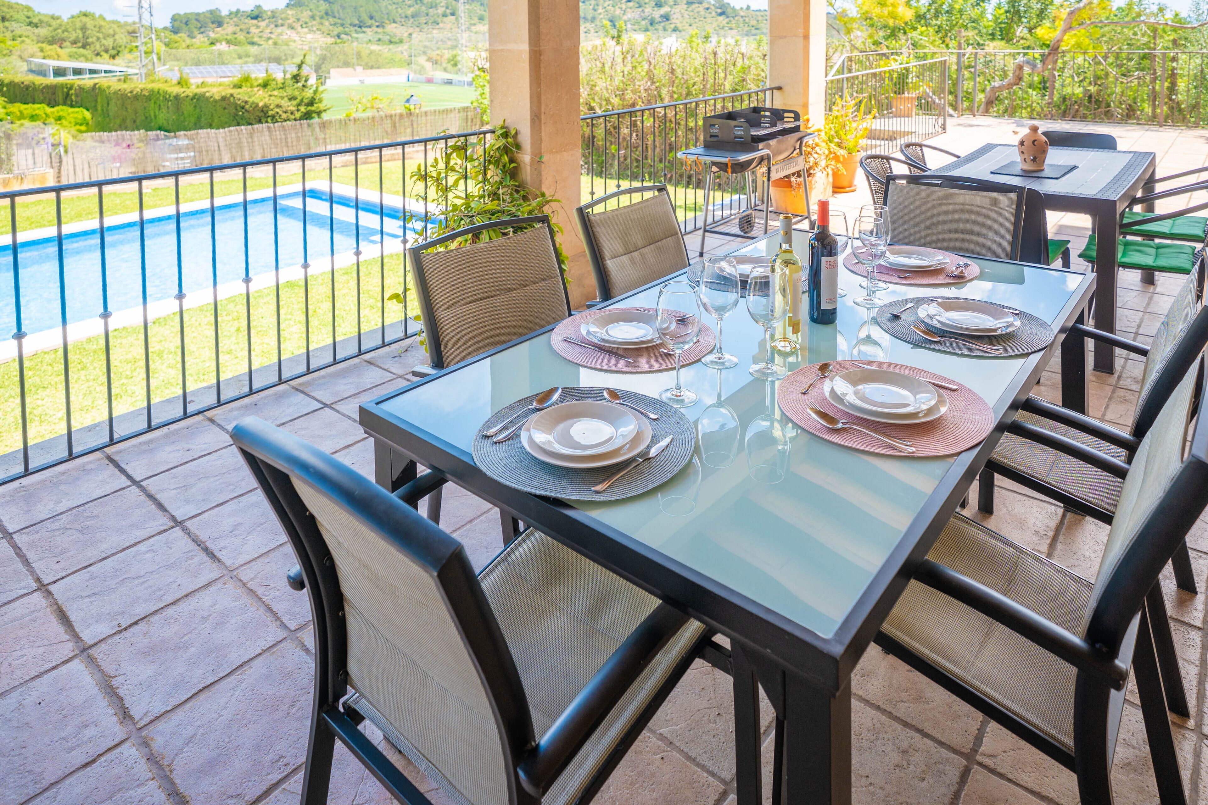 A glass-top dining table set for four with chairs, a pool in the background, and a view of the surrounding landscape.