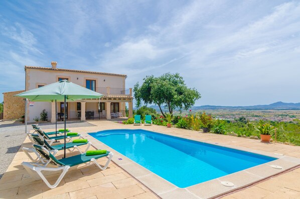 A swimming pool with lounge chairs and a patio area, a building with a balcony, and a view of the landscape.