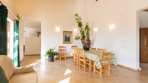 A dining area with a wooden table and chairs, surrounded by potted plants and a painting on the wall.