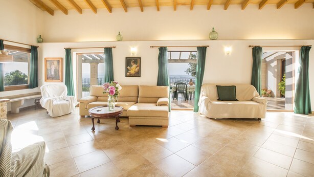 Ein geräumiges Wohnzimmer mit hohen Holzbalken, zwei Sofas, einem Couchtisch und Blick ins Freie durch große Fenster mit grünen Vorhängen.
