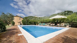 Una piscina rettangolare con bordo bianco, circondata da piastrelle in terracotta. Si vedono lettini e un ombrellone bianco sotto un albero.