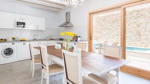 A modern kitchen with a wooden dining table, white chairs, a washing machine, and a stone wall visible through a glass door. - CAN DIANA - Vacation home with private pool in peaceful town. Free WiFi (Campanet)