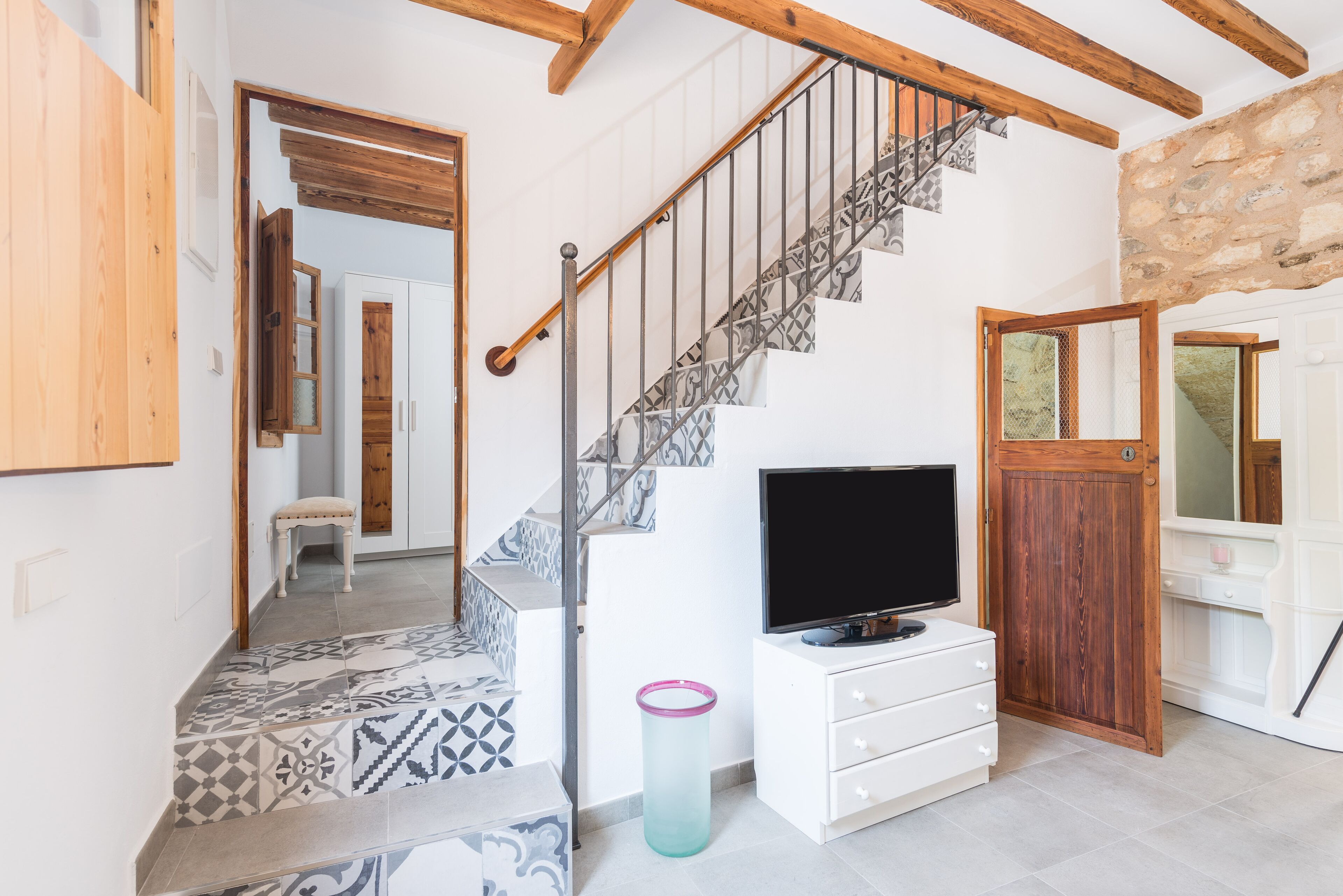 A staircase with patterned tiles, a television on a white dresser, and a stone wall.