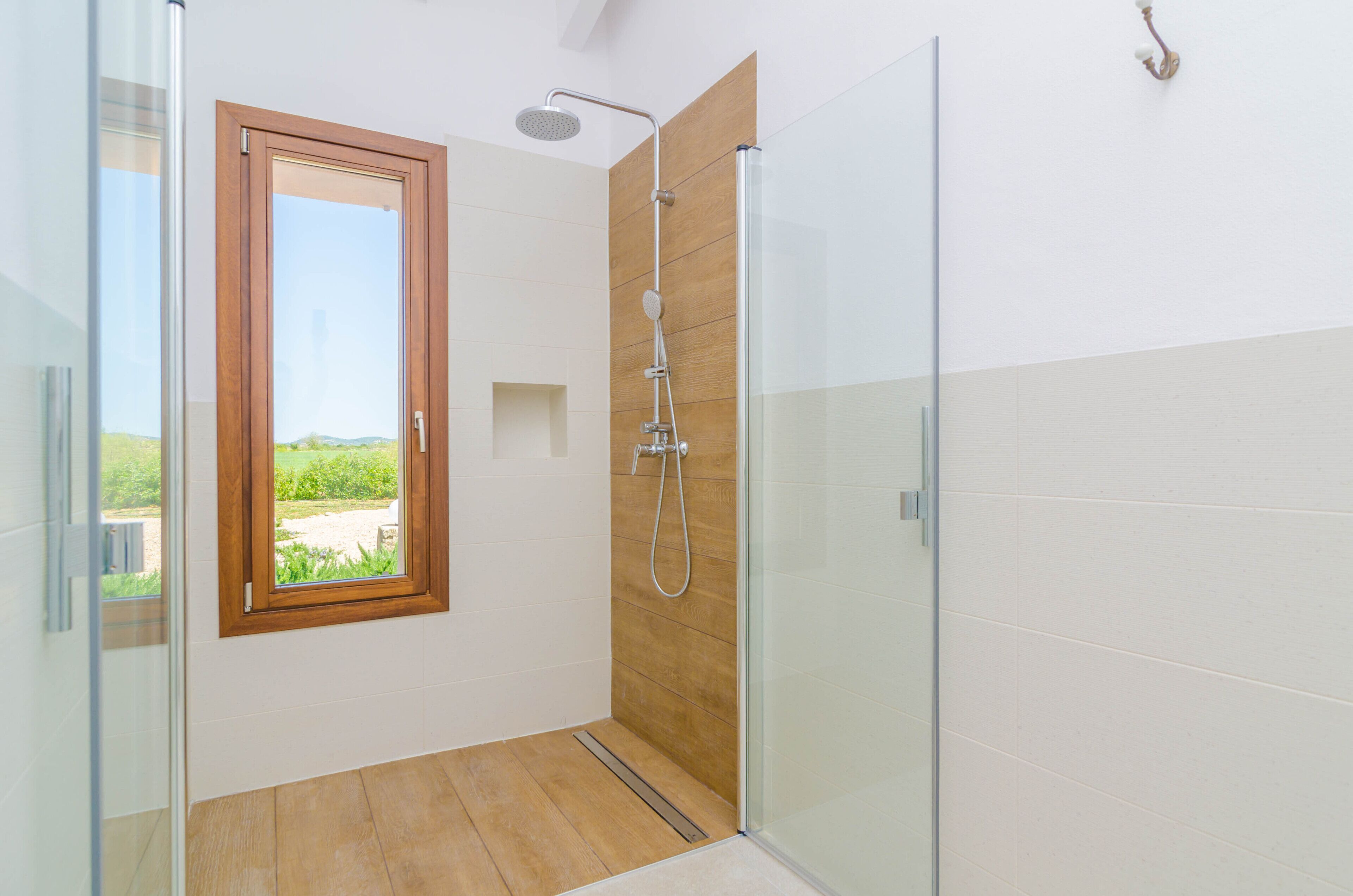 A modern bathroom with a glass shower enclosure, wooden paneling, and a window offering a view of greenery.