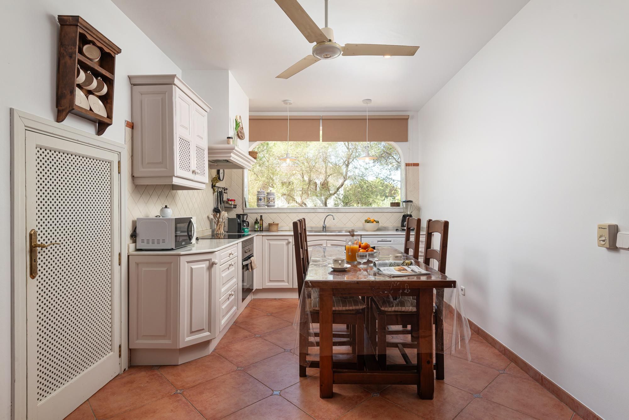 Une cuisine avec des meubles blancs, une table à manger avec des chaises, un ventilateur de plafond et une fenêtre donnant sur des arbres.