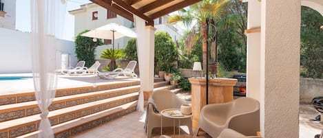 A covered outdoor area with a pool, lounge chairs, and a table with a lamp.