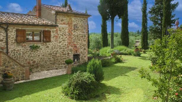 Un bâtiment en pierre avec un toit en tuiles, un petit jardin avec différentes plantes et un sentier.