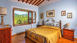 Un dormitorio con cama, mesita de noche, cómoda, ventana con contraventana abierta y cuadros en la pared.