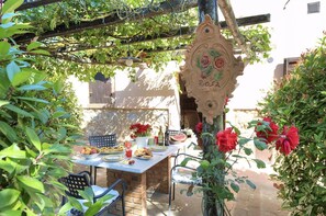 Un espace de restauration en plein air au charme rustique, avec un mur en pierre, une pergola en bois et une table dressée avec de la nourriture et des boissons.