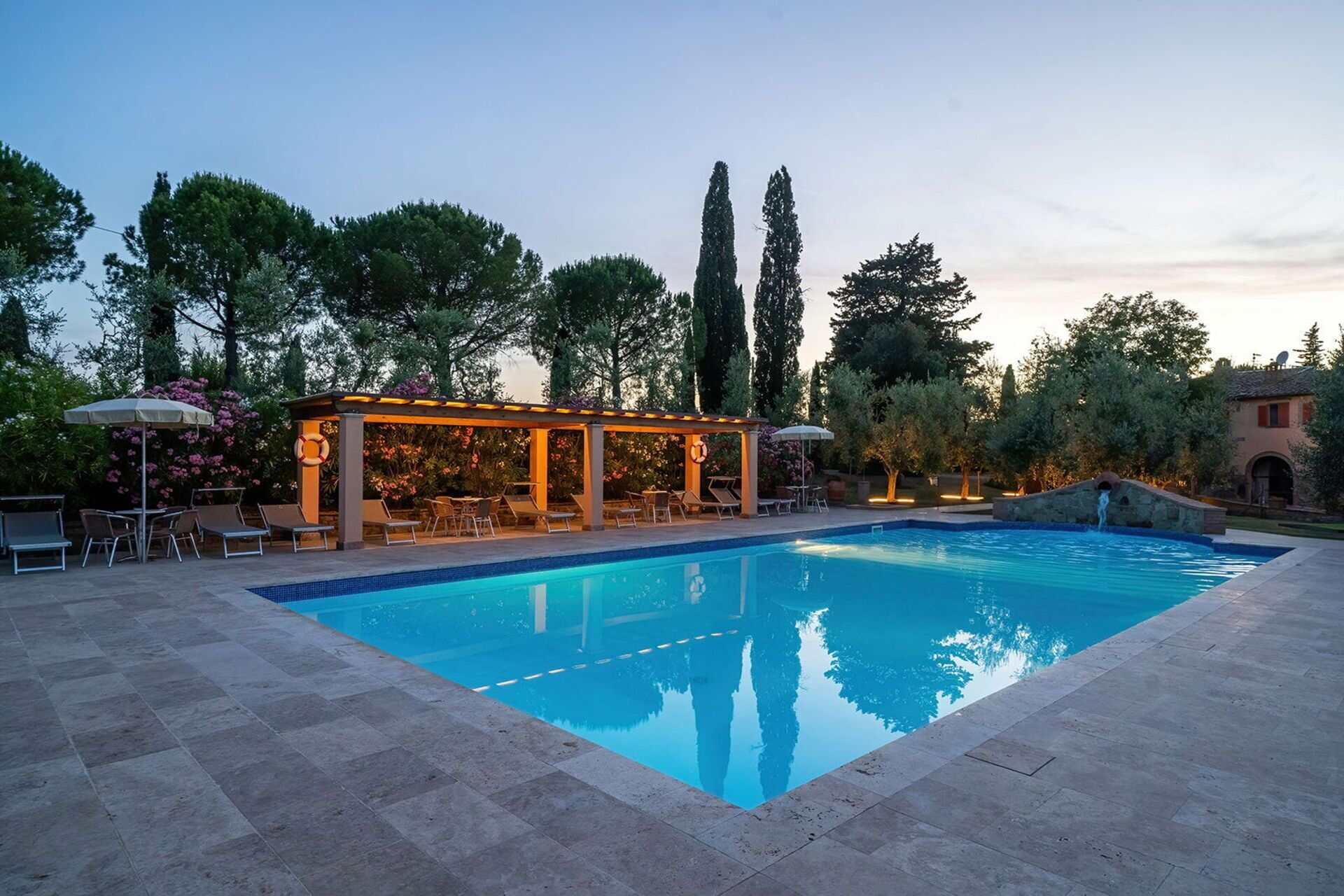 En rektangulär swimmingpool med en pergola och solstolar, omgiven av träd och en stenmur.
