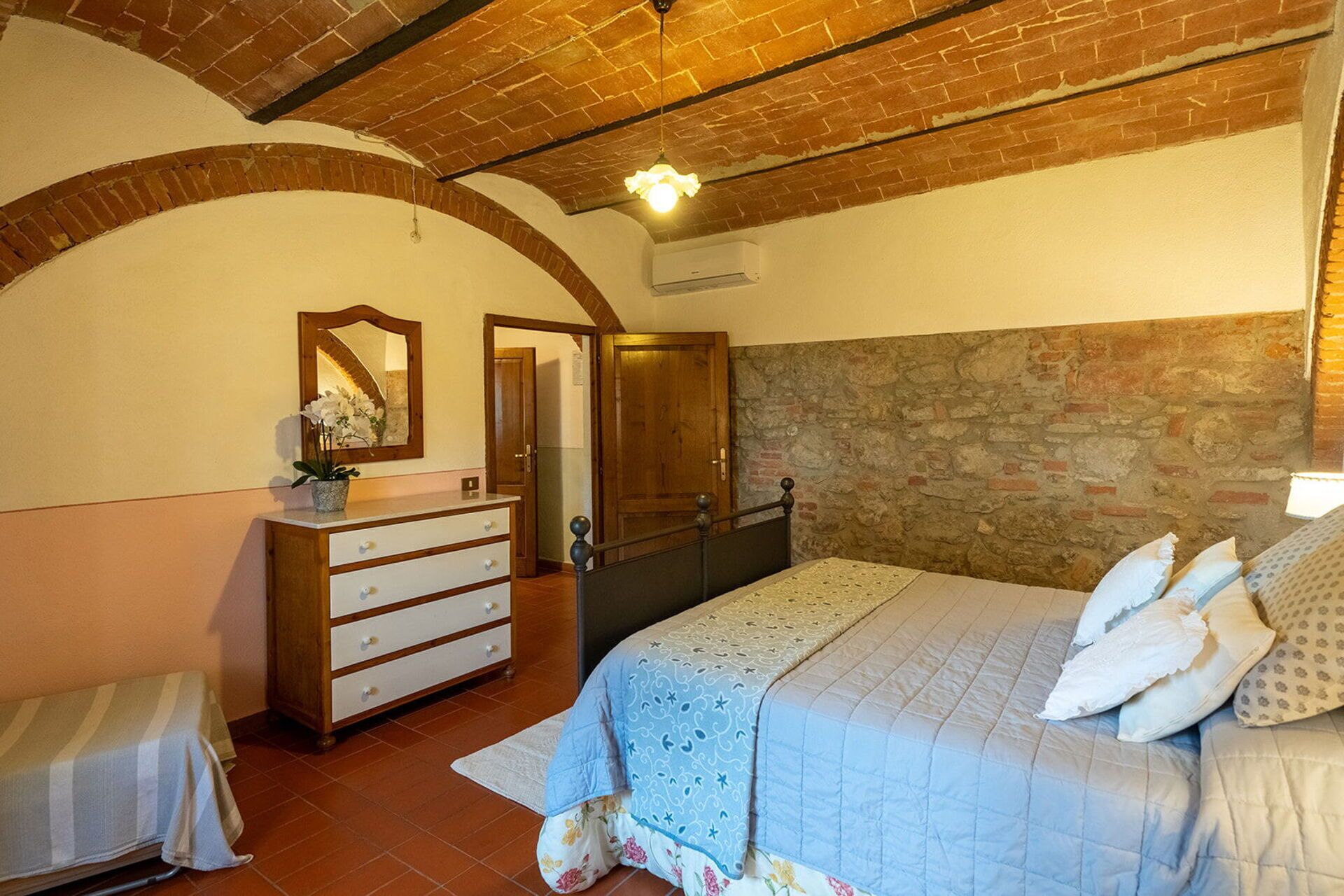 A bedroom with a brick ceiling, a stone wall, a bed with patterned bedding, a dresser, a mirror, and an arched doorway.