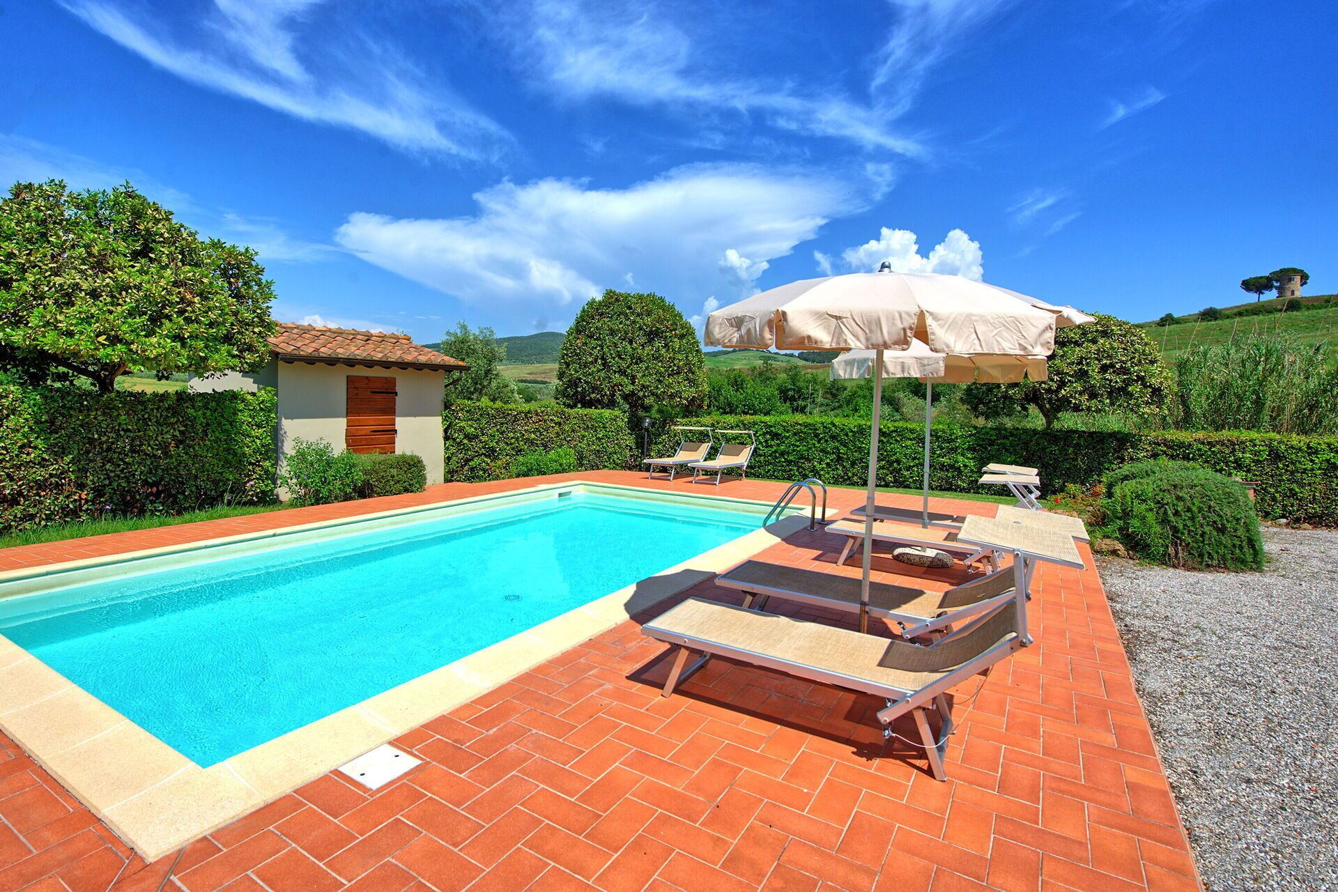 Una piscina rectangular con borde de azulejos, rodeada por un patio de ladrillo. Hay dos sillas de descanso y una sombrilla.