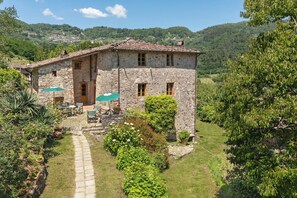 A stone house with a patio, surrounded by lush greenery and hills in the background.