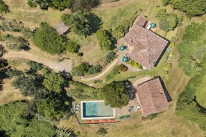 An aerial view of a property with a swimming pool, two buildings with tiled roofs, and surrounding greenery.