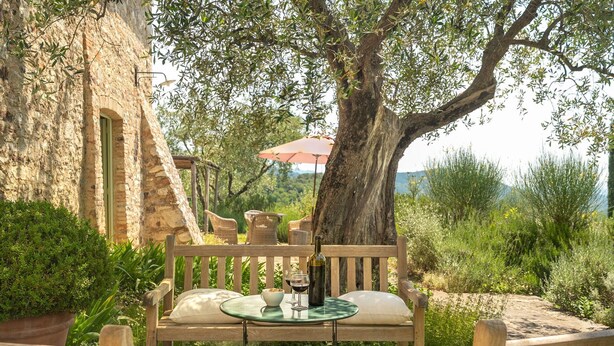 Una panca di legno con cuscini, un tavolo in vetro e una bottiglia di vino, con a vista un muro in pietra, vegetazione sullo sfondo e un ombrellone rosso.