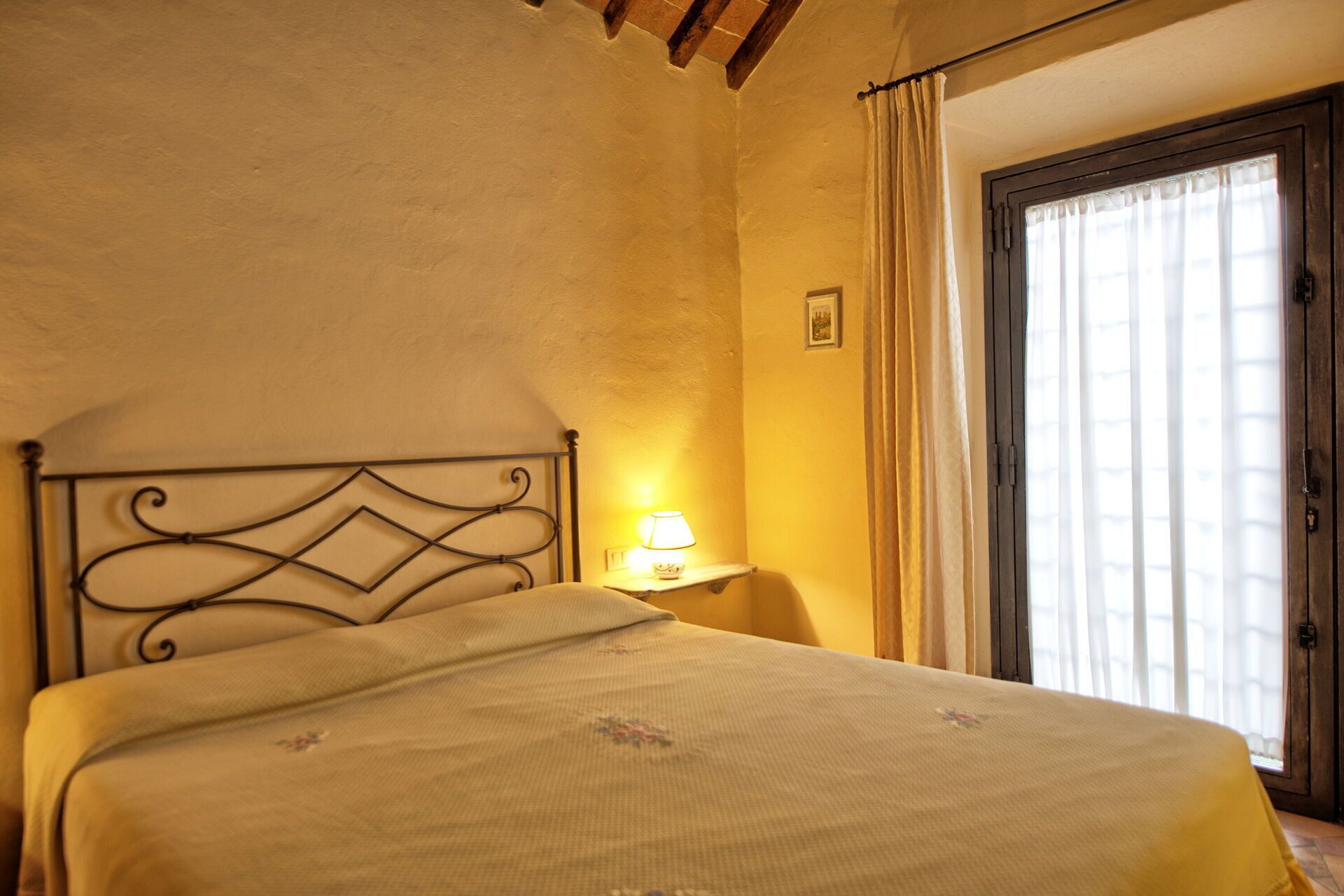 A single bed with a metal headboard in a room with a plain wall and a window with curtains.