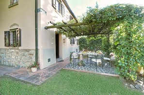 A courtyard with a stone table and chairs, surrounded by greenery and a pergola.
