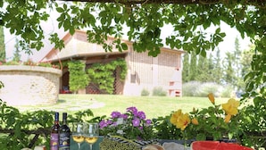 Ein Tisch gedeckt mit Wein, Gläsern, Brot und einer Wassermelone, mit einem Haus und Garten im Hintergrund.