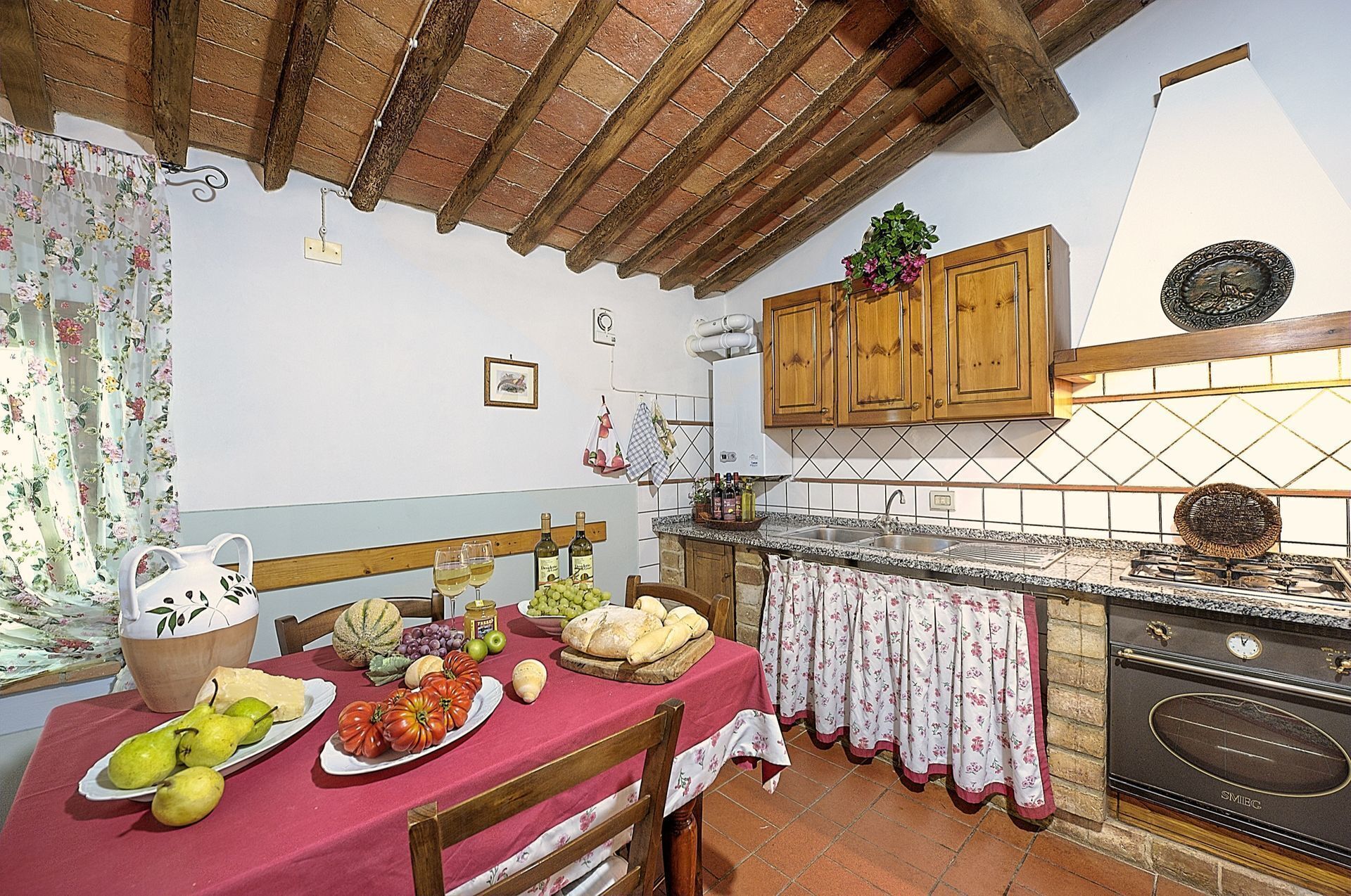 Una cocina rústica con armarios de madera, salpicadero de azulejos y una mesa cubierta con un mantel rojo, dispuesta con frutas y botellas.