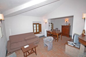 A living room with a brown sofa, a small wooden table, a patterned armchair, a dining table with chairs, and a cabinet.