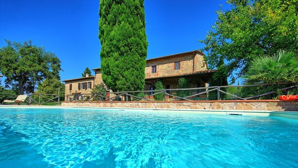Ein Swimmingpool mit klarem, blauem Wasser, umgeben von einer Steinmauer und einem Holzzaun. Im Hintergrund ein Steinhaus mit einem Ziegeldach und einem hohen Baum.
