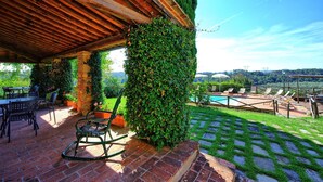 Eine überdachte Terrasse mit Backsteinboden, Sitzgelegenheiten im Freien und Blick auf einen Pool und Gartenlandschaft.