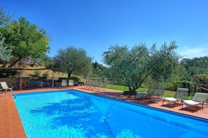 Una piscina rettangolare con acqua cristallina, circondata da un solarium piastrellato. Sono presenti diverse sedie a sdraio e un tavolino con sedie. L'area è ombreggiata da alberi e sullo sfondo si vede un muro in pietra.