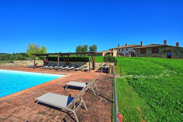 A poolside area with lounge chairs and a pergola, a stone house with a tiled roof, and a grassy hill in the background.