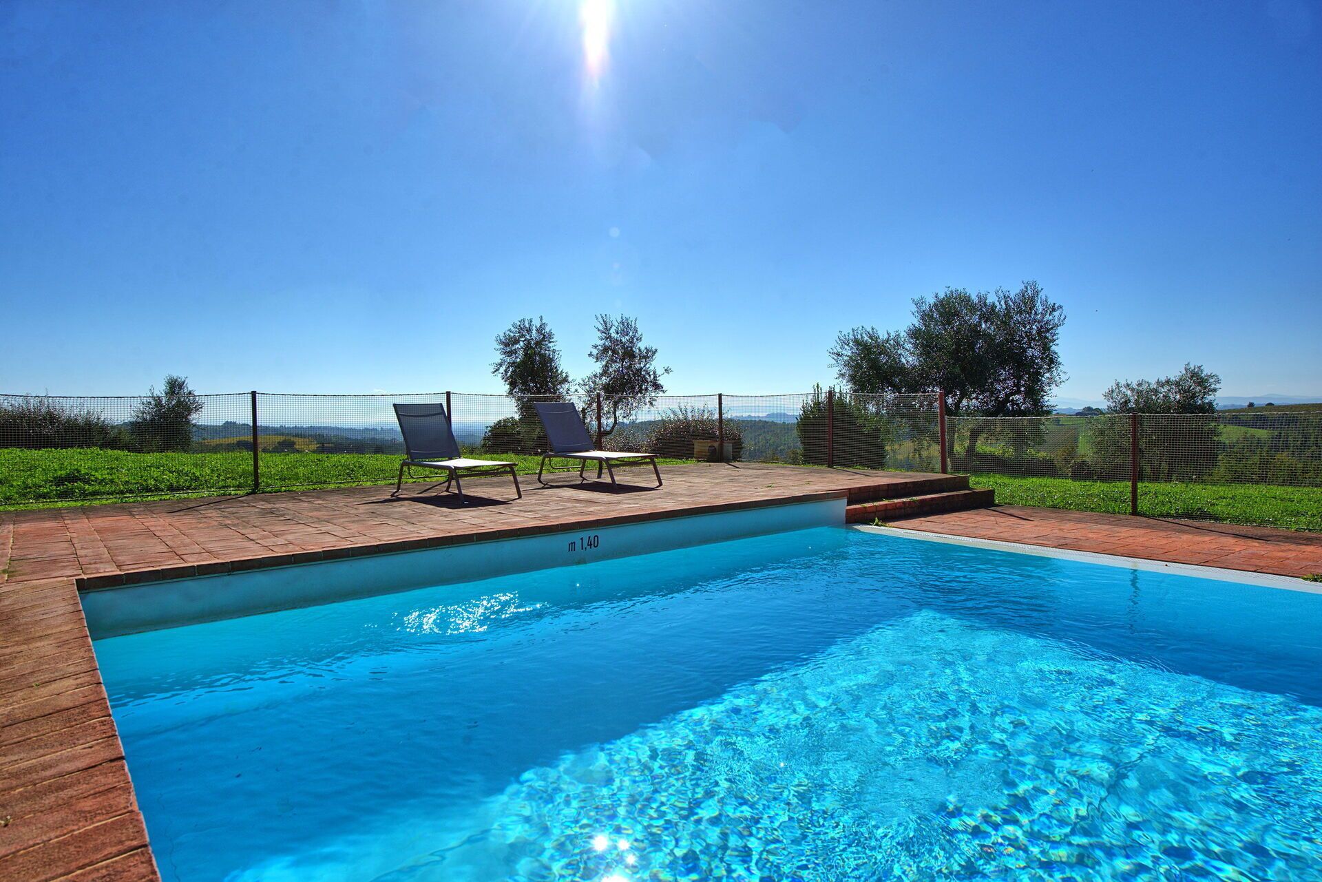 Una piscina rectangular con superficie de agua azul cristalina, rodeada por un deck de ladrillo. Hay dos sillones de descanso y una mesita en el deck.