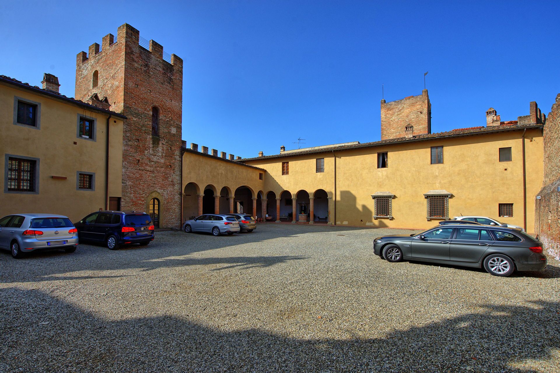 A courtyard with a historic building featuring a tower and arches, surrounded by parked cars.