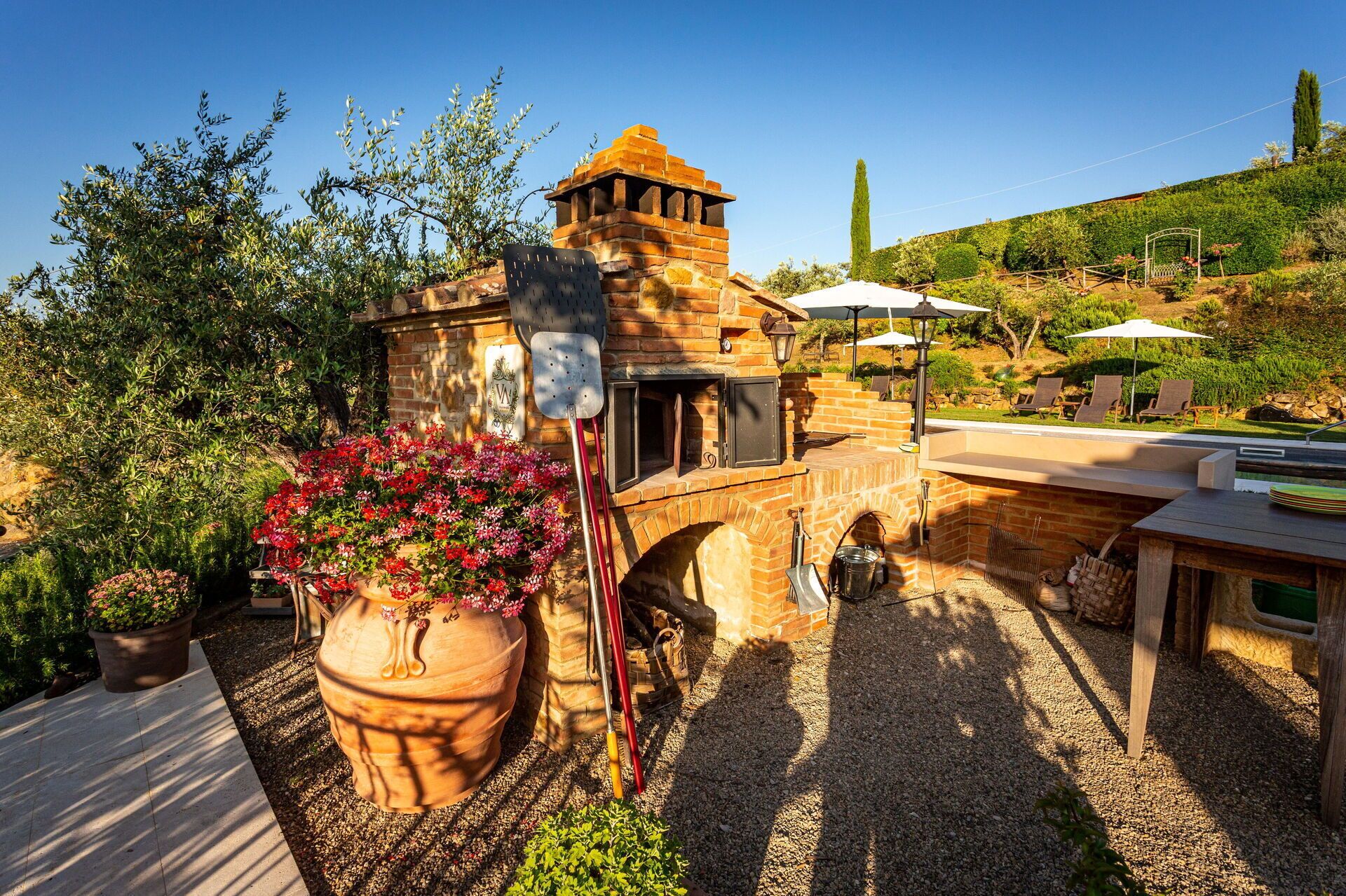 Una cocina exterior de ladrillo con una parrilla, una maceta grande y un patio con sillones y sombrillas.