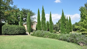Un jardin bien entretenu, agrémenté de grands cyprès, d’un bâtiment en pierre et de diverses espèces d’arbustes.