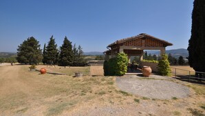 Un área de estar rústica al aire libre con una pérgola, rodeada por un campo seco y árboles.