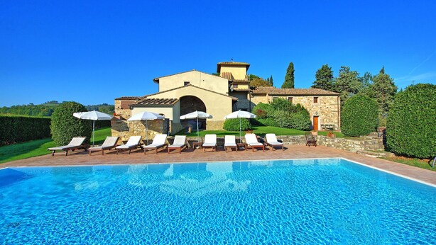 Zona de piscina con sillones y sombrillas, un edificio de piedra con tejado de baldosas y un cielo azul despejado.