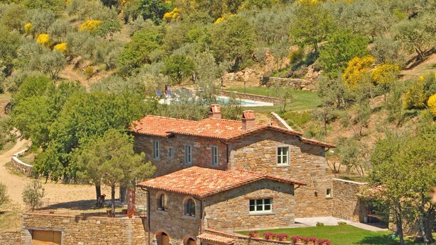 A stone house with a red-tiled roof, surrounded by a garden and trees.