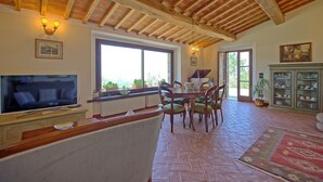 A spacious living area with a large window, wooden ceiling beams, a dining table with chairs, a television, and a cabinet with glass doors.
